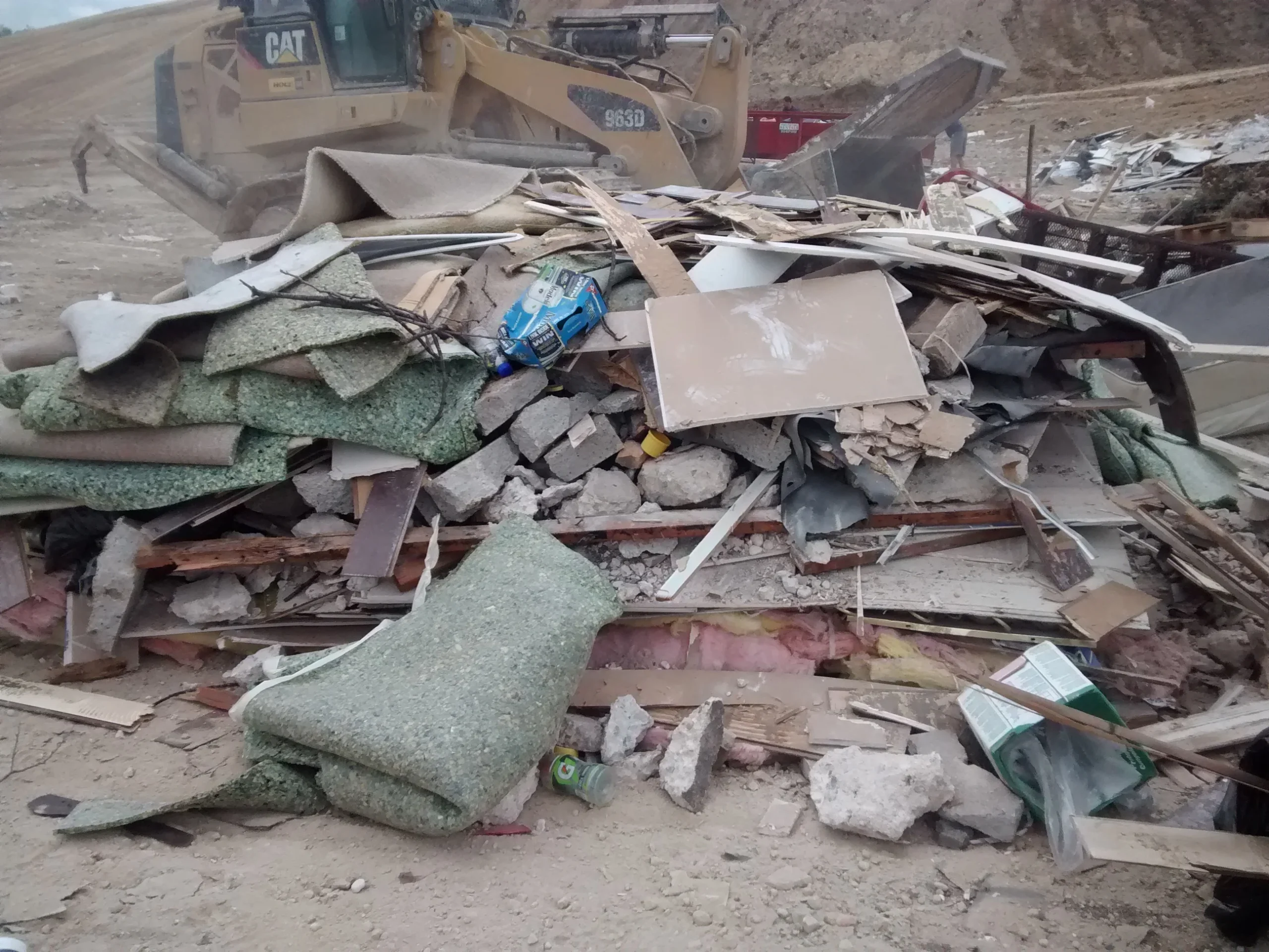 A pile of construction debris, including concrete, wood, carpeting, and other materials, at a construction site with heavy machinery in the background.