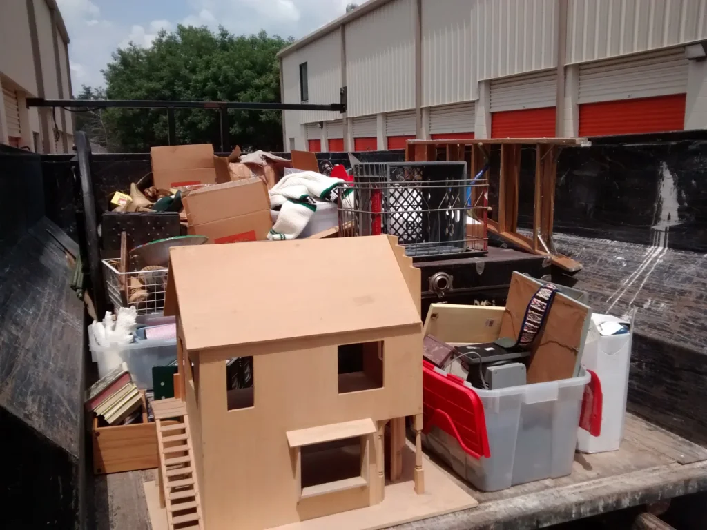 A large dumpster filled with various items, including boxes, a wooden dollhouse, furniture, and household goods, located near storage units with orange doors.