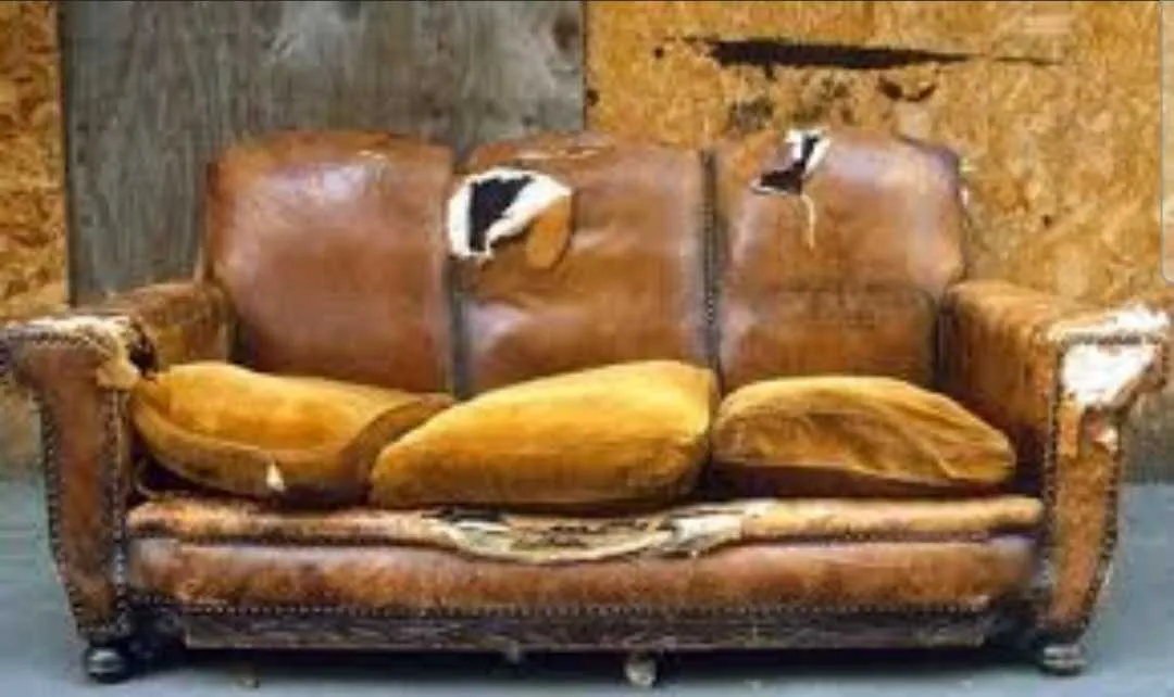 A worn-out brown leather sofa with torn cushions and visible damage, set against a wooden background.