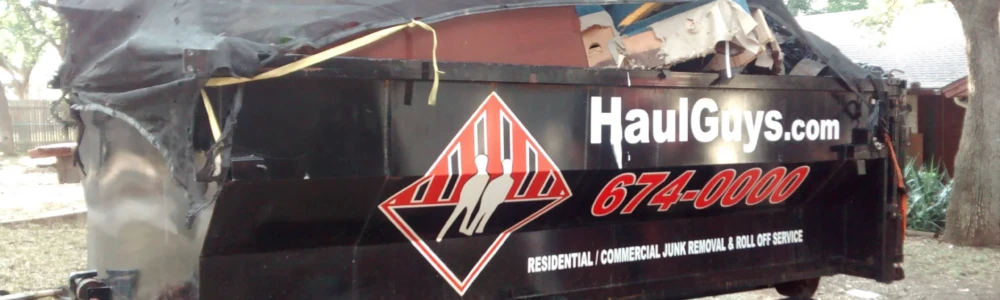 A Haul Guys trailer loaded with various junk materials and covered by a black tarp, featuring the company's logo and contact number, parked in a residential area surrounded by trees.