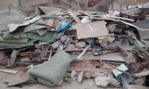 A pile of construction debris, including concrete, wood, carpeting, and other materials, at a construction site with heavy machinery in the background.